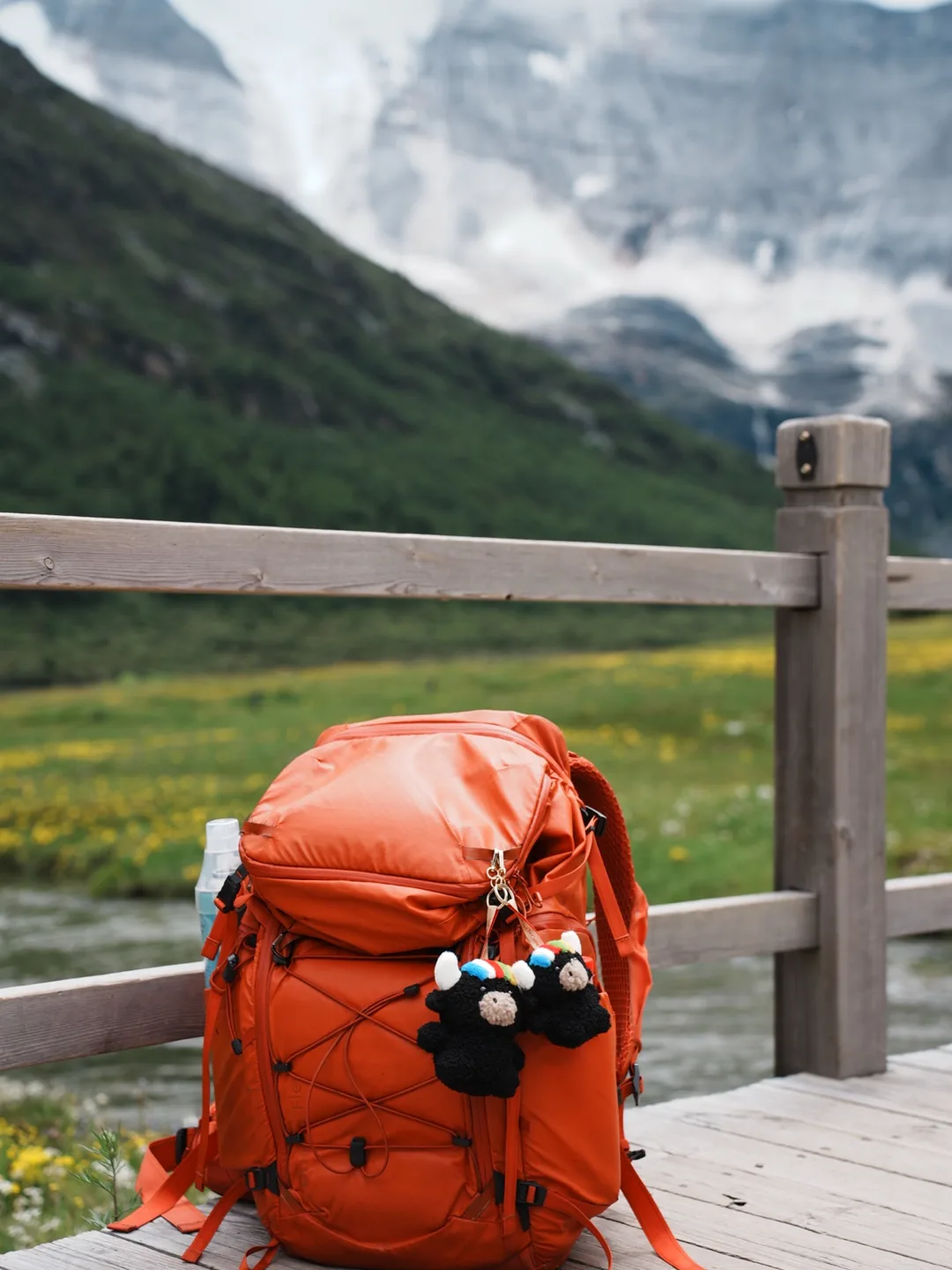 Two Tashi yak charms hanging on an orange hiking backpack in front of snow mountains and a green meadow — 雪山草甸前橙色背包上的扎西掛件