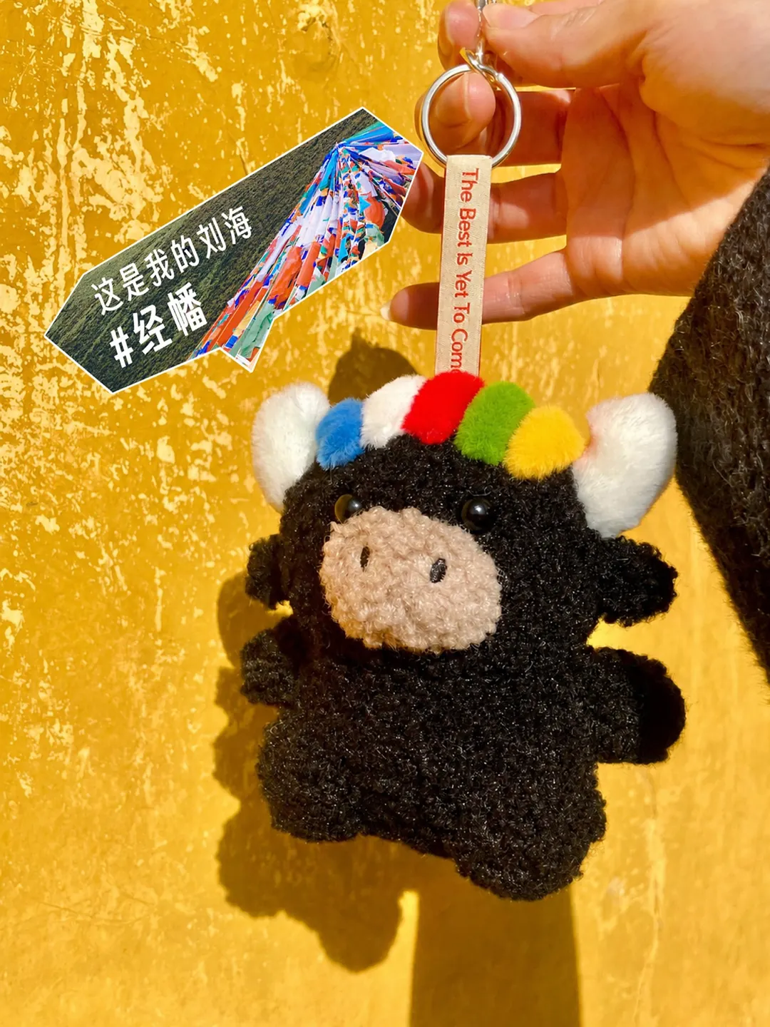 Close-up of the Tashi keychain held against a yellow wall, its five-colour fringe echoing the prayer flags — 黃牆前的扎西掛件特寫，五色經幡劉海